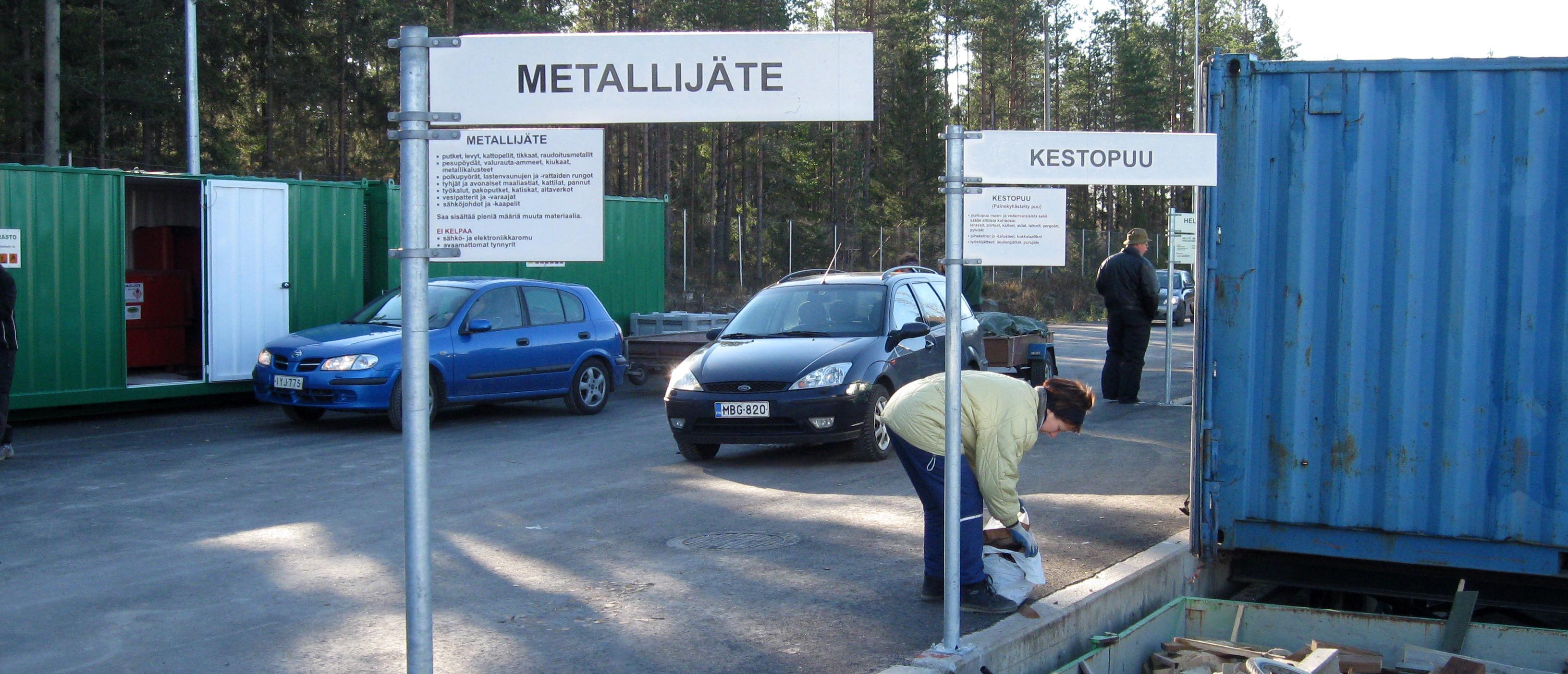 Kuva Hollolan jäteasemalta, missä asiakas purkaa jätteitään metallilavalle ja autoja on pysäköitynä asfaltoidulle kentälle.