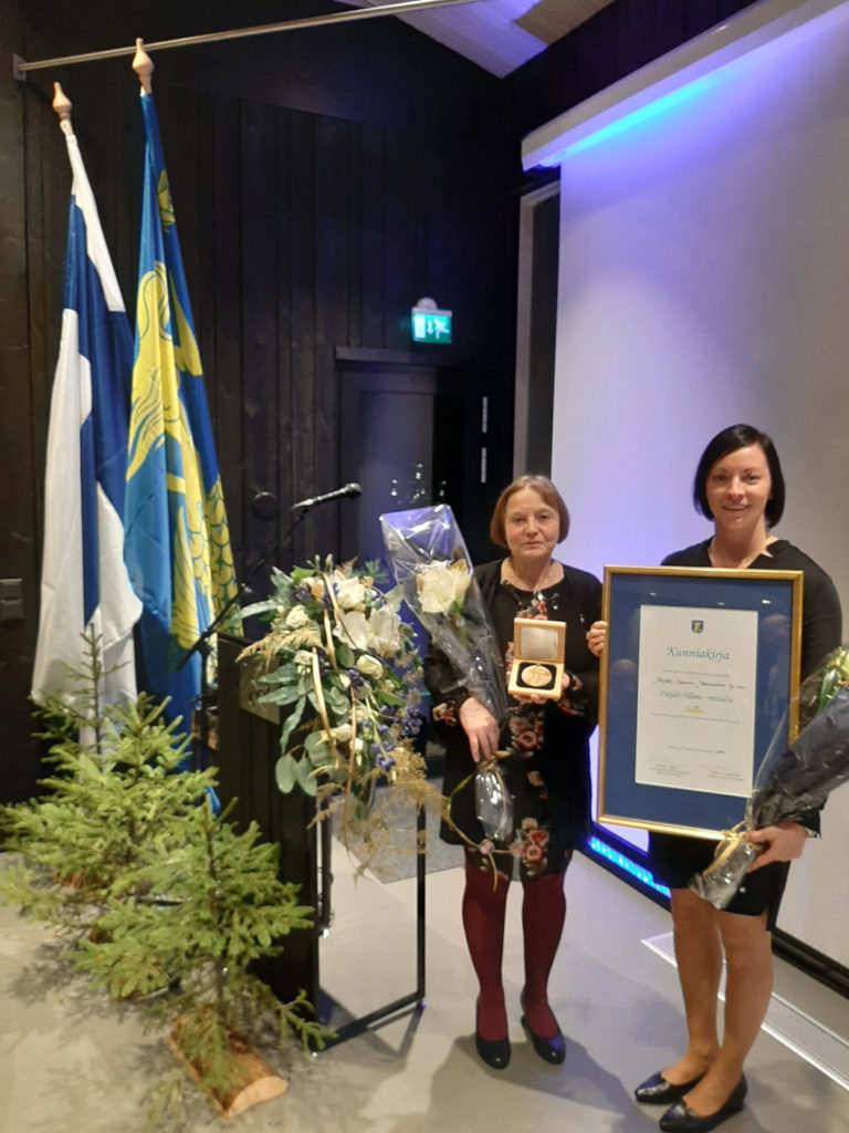 Tuula Honkanen ja Johanna Rusanen kuvattuna Wellamo-juhlassa pidellen vastaanotettua palkintoa, kunniakirjaa ja kukkakimppua.