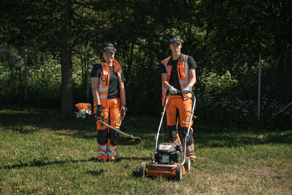 kaksi ihmistä oransseissa työvaatteissa seisoo nurmikolla puiden edessä
