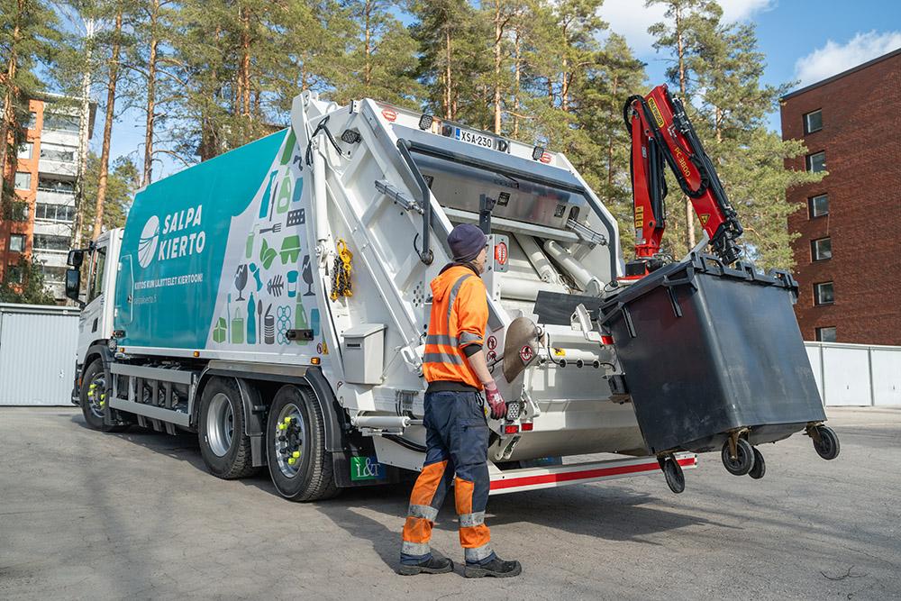 oranssipukuinen jätekuljettaja tyhjentää harmaan sekajäteastian sisällön jäteautoon
